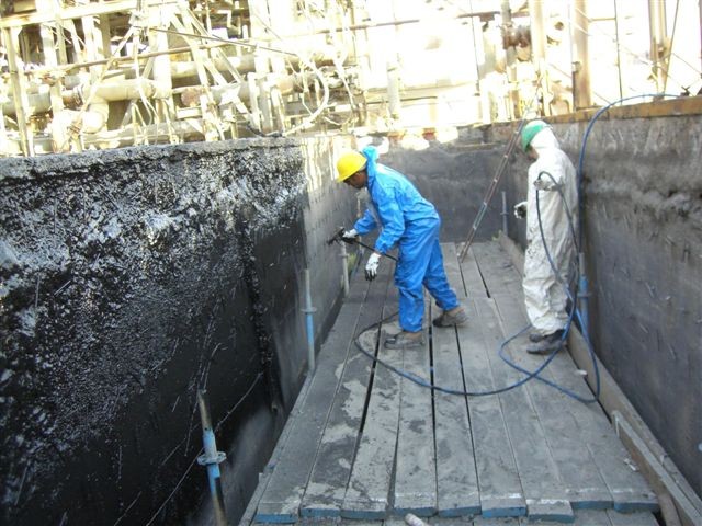 Two workers wearing protective suits and helmets are applying a coating or spray treatment to the interior walls of a large industrial concrete tank or containment structure.