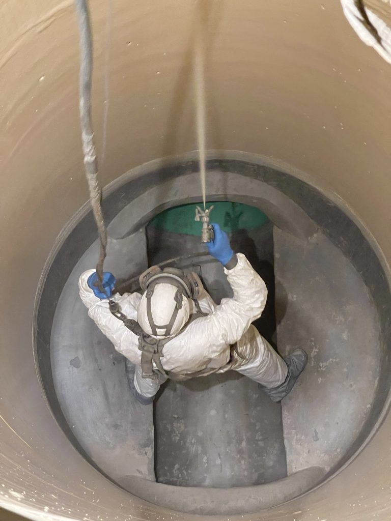 A worker in protective gear is inside a confined chamber while applying a protective coating.