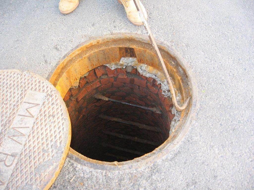 This photo shows an open manhole in a paved surface, with the metal cover pushed aside. Inside, the structure appears to be constructed of brick, with visible deterioration and missing mortar along the upper interior wall.