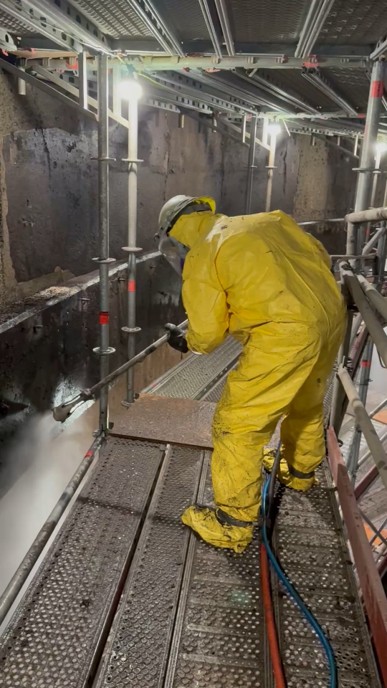 A worker in full protective gear performs high-pressure surface preparation inside a wastewater structure.