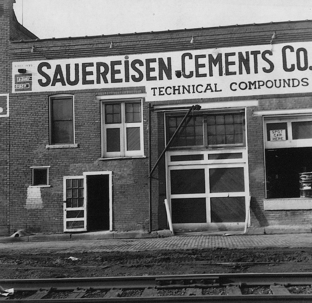 This black and white photo shows an older brick industrial building with a large "Sauereisen Cements CO." sign on the front.
