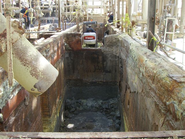 This photo shows a deteriorated sulfur pit with heavy corrosion and staining, while workers and equipment are positioned around it inside a processing facility.