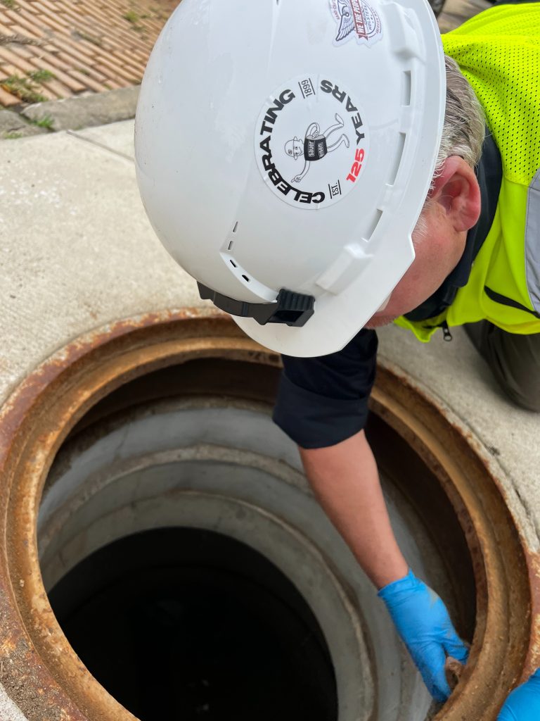 This photo shows a worker positioned over a manhole opening, preparing and performing work at the surface while wearing safety gear. 