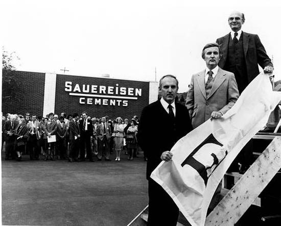 A black-and-white photo shows a group of men standing on a platform holding a flag during a formal ceremony, with a crowd gathered behind them in front of the Sauereisen Cements building.