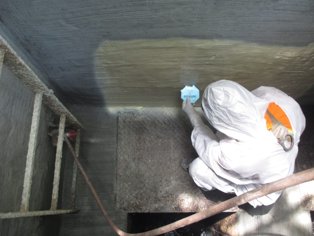 A worker in protective gear is inside a concrete chamber, doing a mil check to the wall by hand while kneeling.