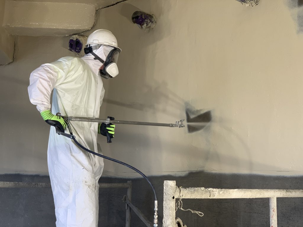 A worker wearing protective gear using a spray gun to apply a coating to the walls of a concrete structure.