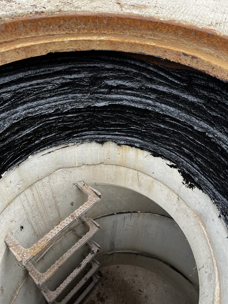 This photo shows the interior of a concrete manhole with a ladder, where a thick black F-88 application has been applied along the upper wall, providing a protective barrier at the crown area.