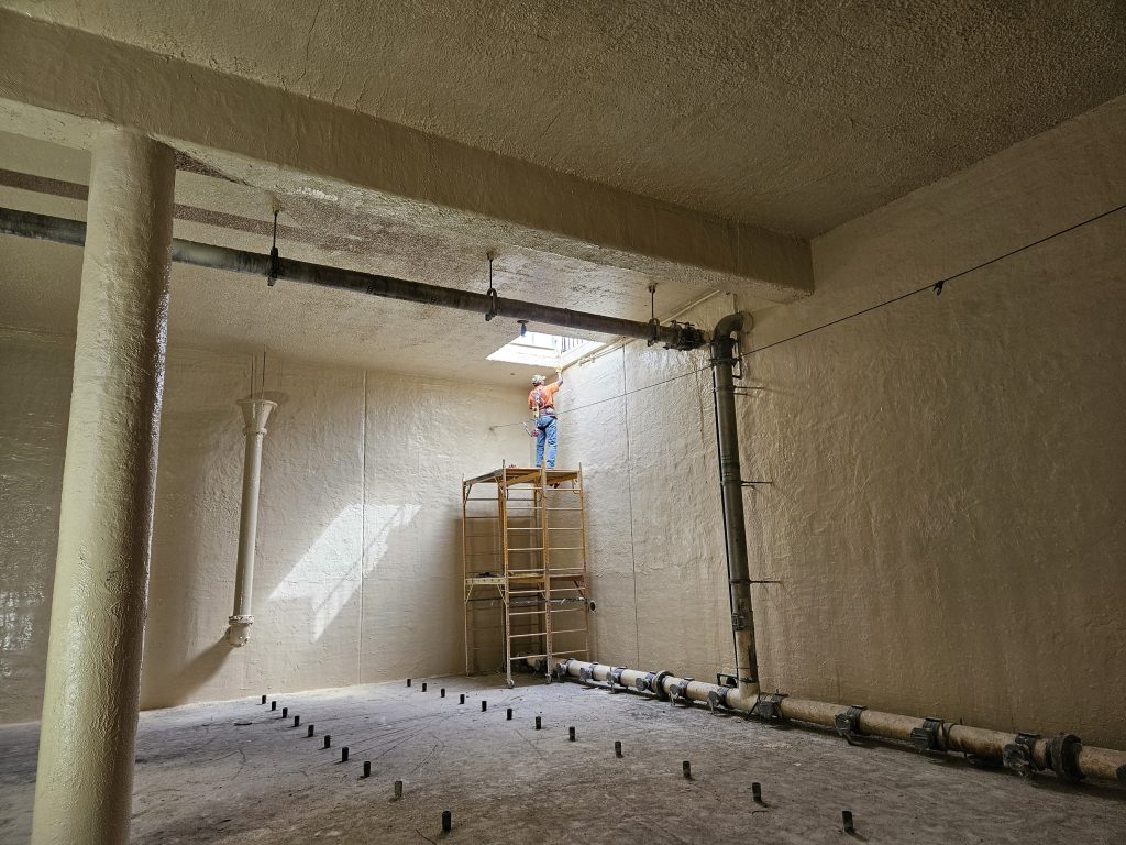 A worker stands on scaffolding inside a large industrial concrete chamber, applying a protective coating to the ceiling and walls.