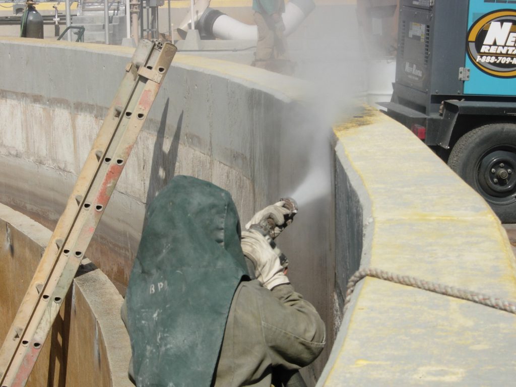 A worker wearing protective gear spraying inside a concrete wastewater structure during surface preparation