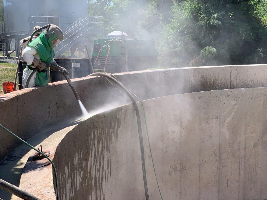 A worker using high-pressure water jetting to prepare the concrete surface of a wastewater tank, removing contaminants and creating the proper profile for a durable protective coating application.